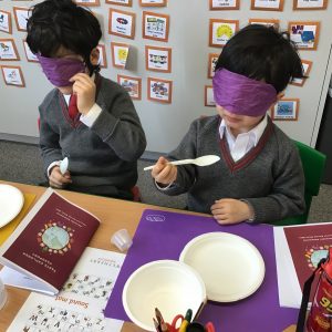 Two kids sat at a desk with plastic spoons, wearing purple blindfolds